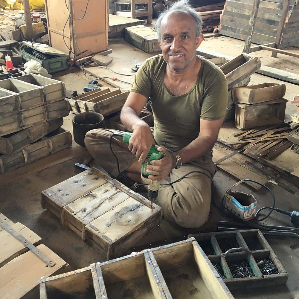 Mand arbejder med genbrugstræ og bruger en elektrisk boremaskine til at samle rustikke trækasser i et værksted fyldt med genbrugsmaterialer og genbrugsmøbler.